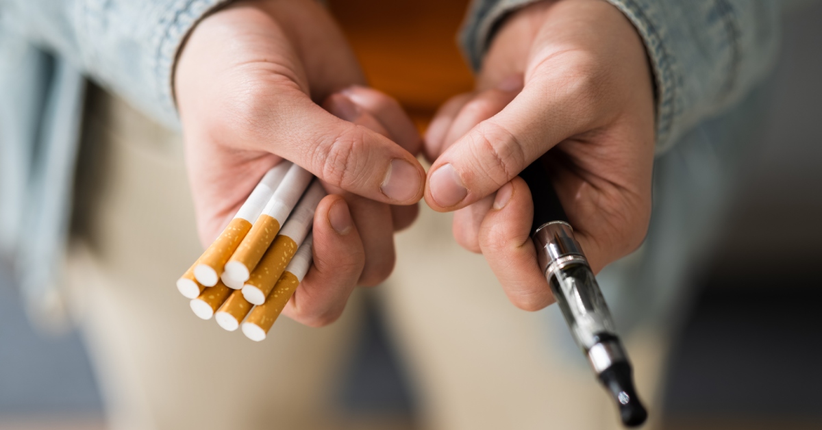 A person holding eight cigarettes in their right hand and a vape pen in their left hand. They wear a denim shirt.