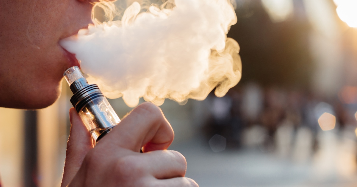 A person holding a vape pen in their hand while blowing out the vapor from their mouth. It appears to be a sunny day.