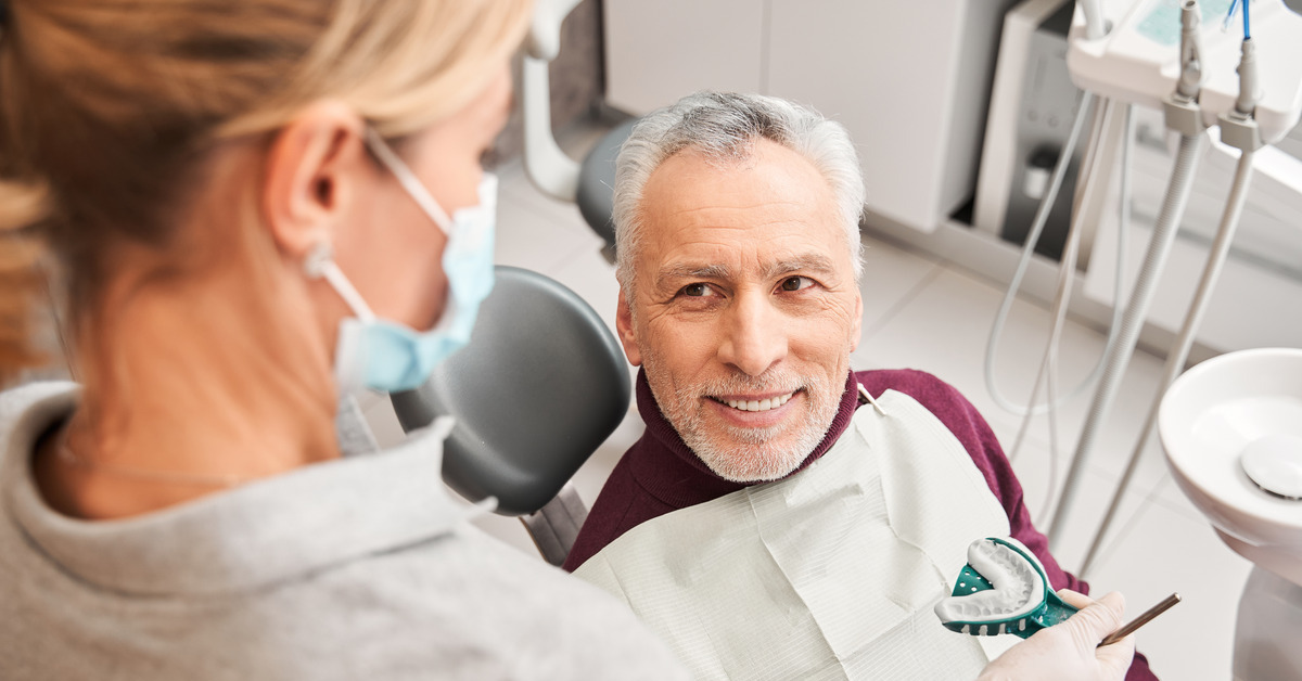 A dentist sits and talks with an older patient while holding dentures. The patient wears a gray shirt.