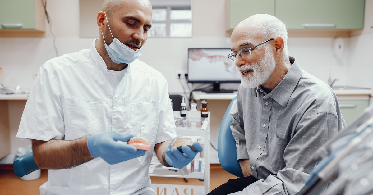 A dentist holds a mouth impression for dentures in front of a patient who smiles at her. The patient wears a turtleneck.