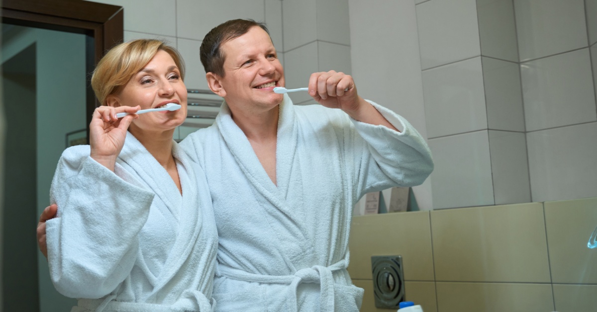 A man and a woman stand in a bathroom brushing their teeth together. They are wearing matching robes.