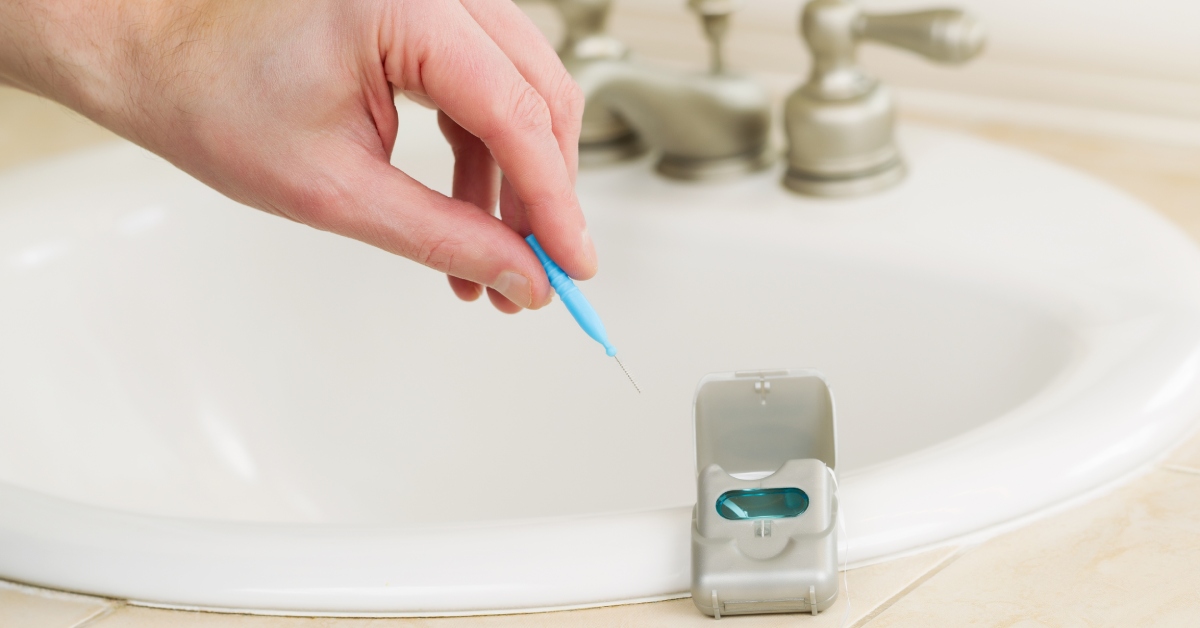 A hand takes a blue dental pick out of a container. The container lies on the edge of a white sink.