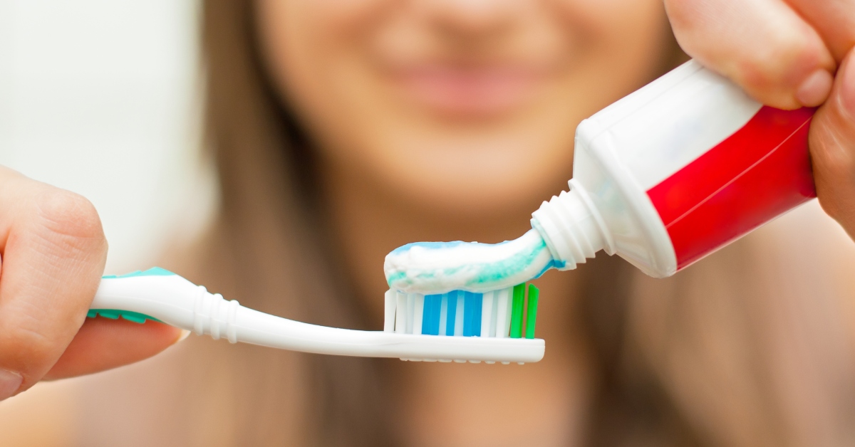 A woman squeezes blue and white toothpaste onto her toothbrush. The toothpaste tube is red and white.