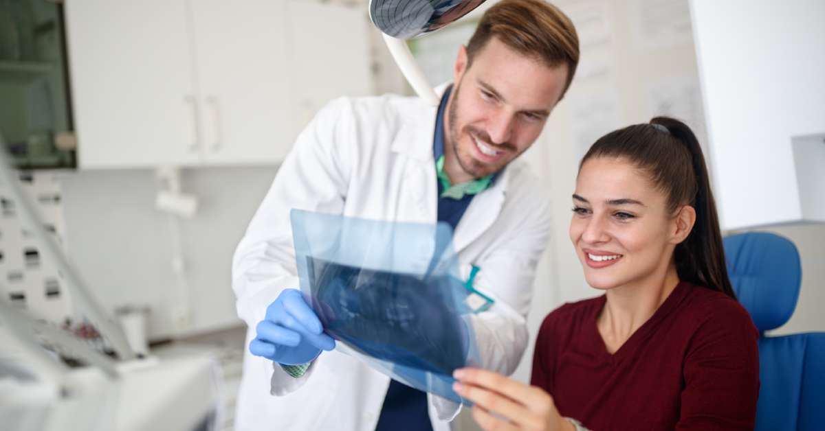 A patient looks at her X-ray with her dentist, who is wearing a white coat and blue gloves. They are in an examination room.