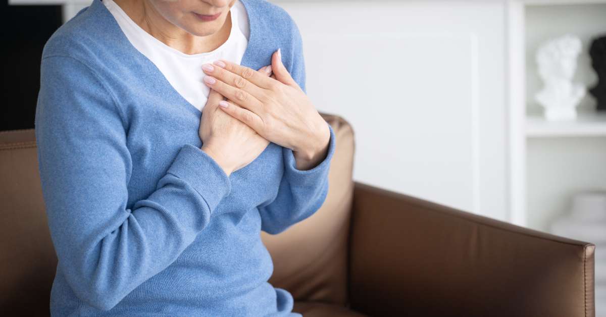 A woman in a blue sweater holds her hands to her chest while sitting in a chair. Her face is pained.