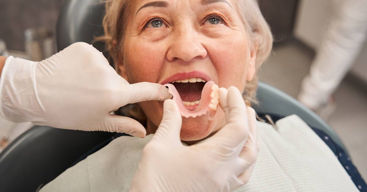 A dentist's hands hold a denture in front of a woman's open mouth. The denture appears to replace only four teeth.