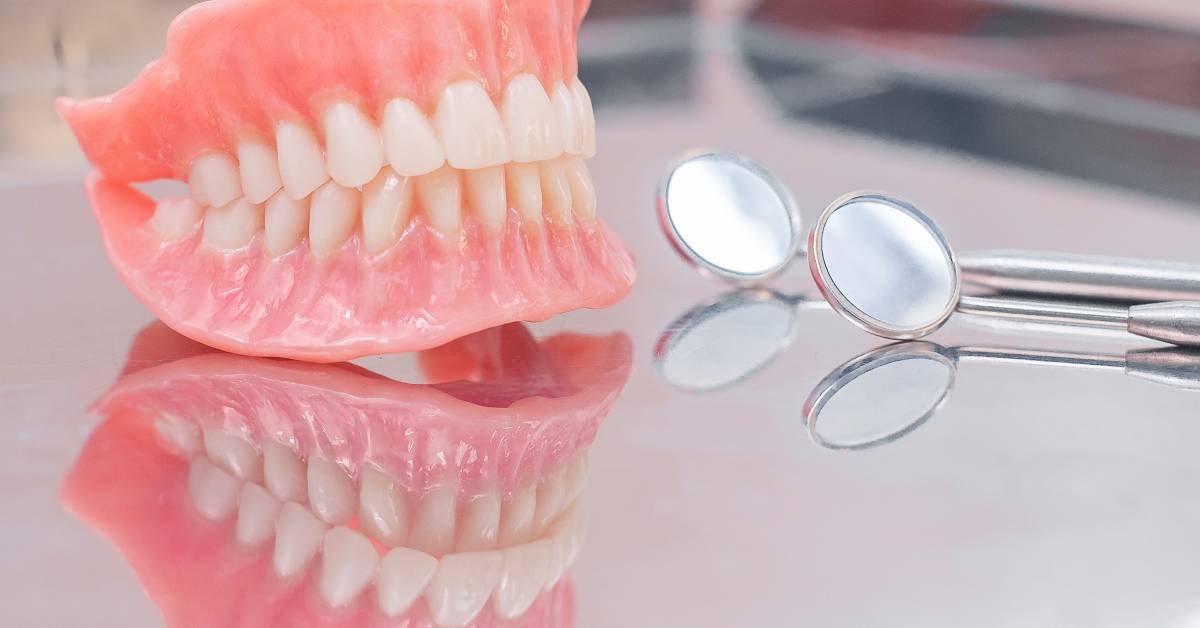 A pair of dentures sits on a silver counter next to a pair of dental mirrors. The metal reflects all three items.