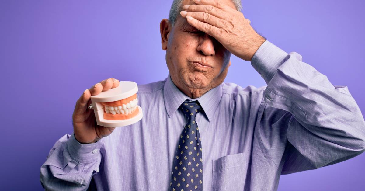 A man with a pained face and his hand on his forehead holds a pair of dentures in his hand. He wears a shirt and tie.