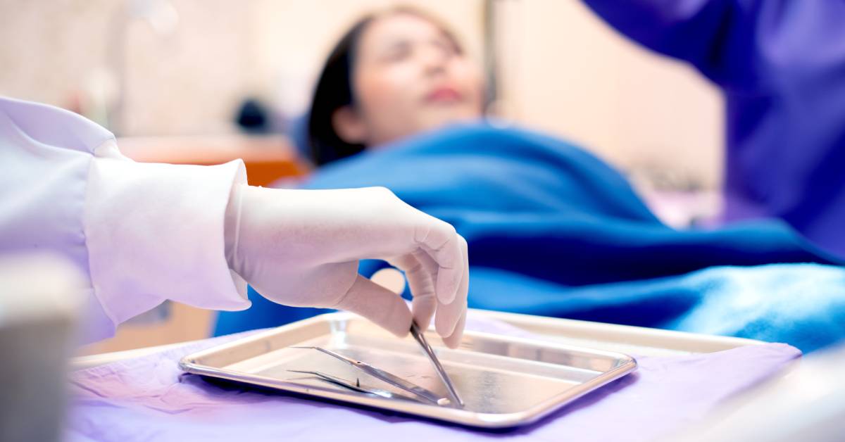 A dentist's hand reaches for silver tools on a table while a patient sits back on a dentist's chair in the background.