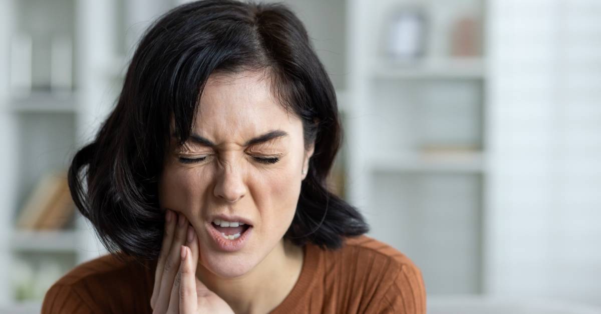 A woman holds her cheek as she experiences a toothache. A bookshelf is behind her in the background.