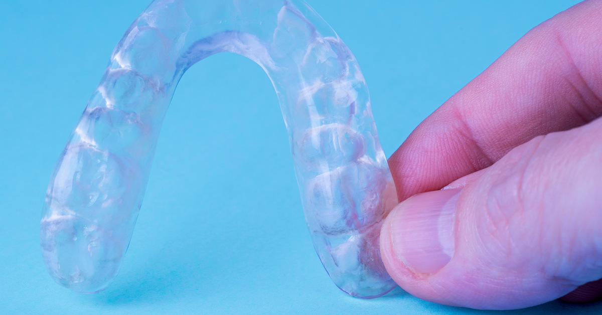 Close-up of a pair of fingers holding a clear plastic aligner for teeth. The aligner sits against a blue background.