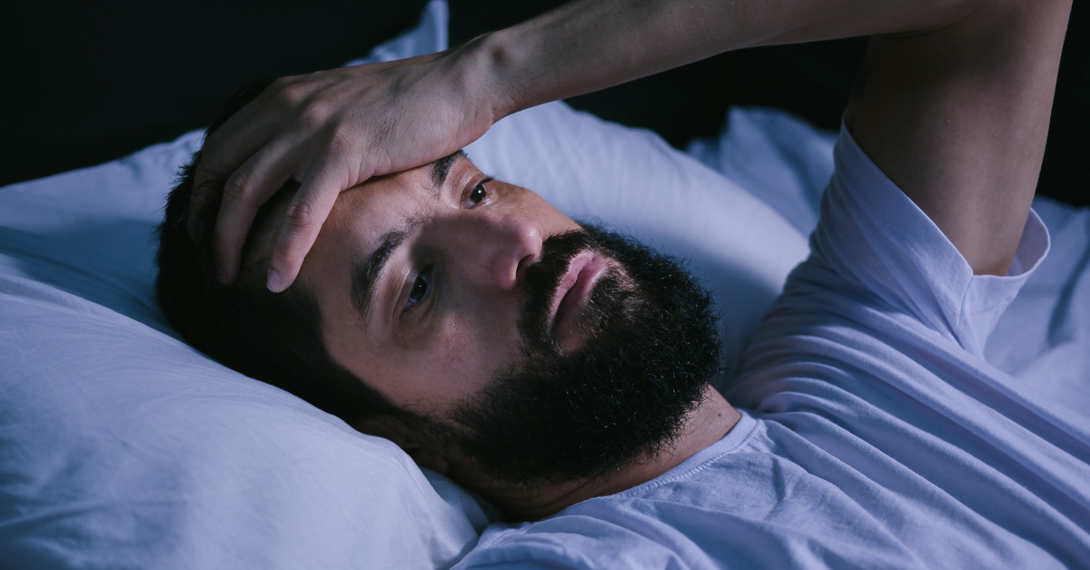 A man lies in bed, looking exhausted. He has his hand resting on his forehead and his head on his pillow.