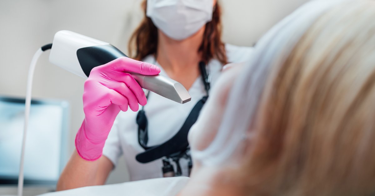 A dentist prepares to insert a scanner into a patient's mouth. She wears a white mask and pink gloves.