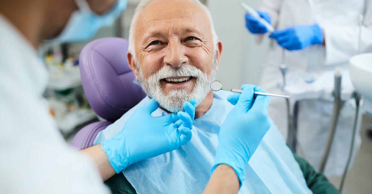 A patient with a white beard smiles as he sits in a dental chair. He wears a blue smock over his shirt.