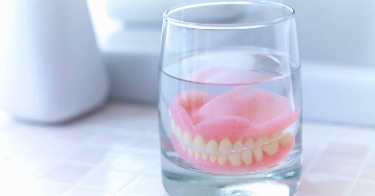 A pair of dentures sit in a glass of water on a white counter. A container of toothbrushes is nearby.