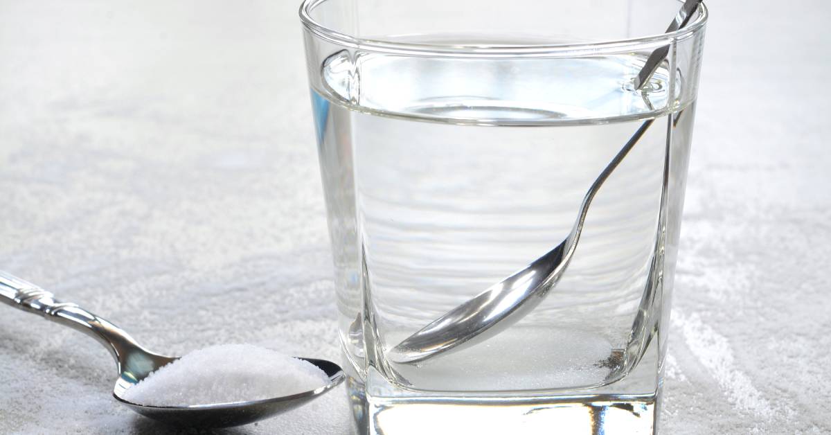 A teaspoon filled with salt sits next to a glass of water on a counter. A spoon is inside the glass.